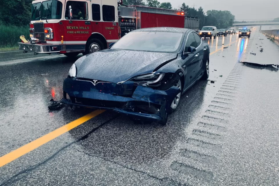 Dark blue Tesla with crumpled front bumper on rainy highway with red fire truck in background